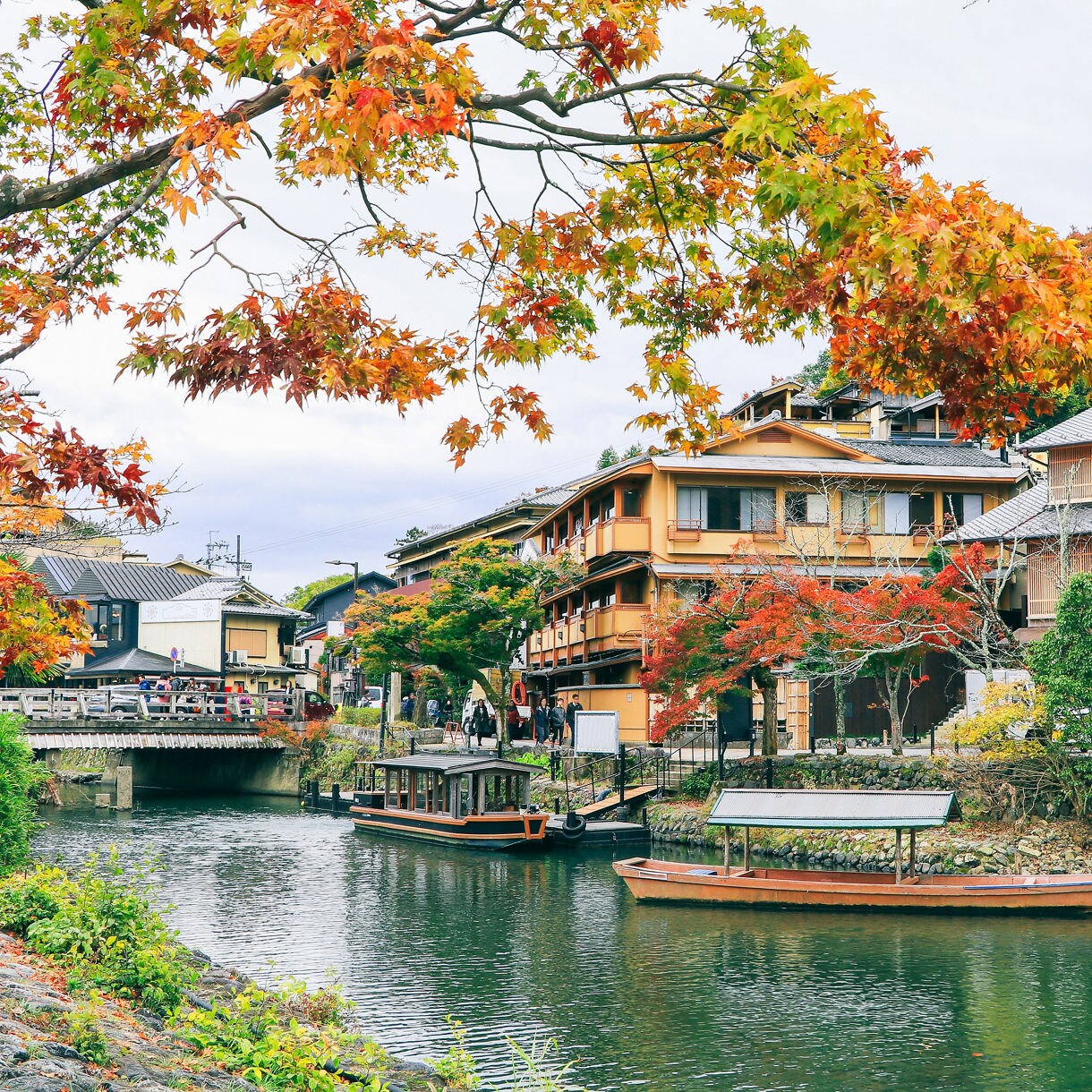 A peaceful canal scene in Kyoto’s Arashiyama district, with wooden boats docked along the water, traditional buildings lining the shore and vibrant autumn leaves hanging overhead.